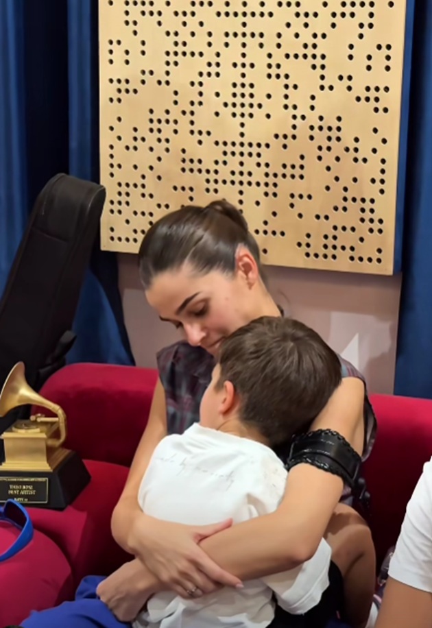 Theo Rose st&acirc;nd pe un scaun și țin&acirc;ndu-l &icirc;n brațe pe fiul ei, Sasha, care stă cu spatele la cameră și &icirc;și ascunde chipul