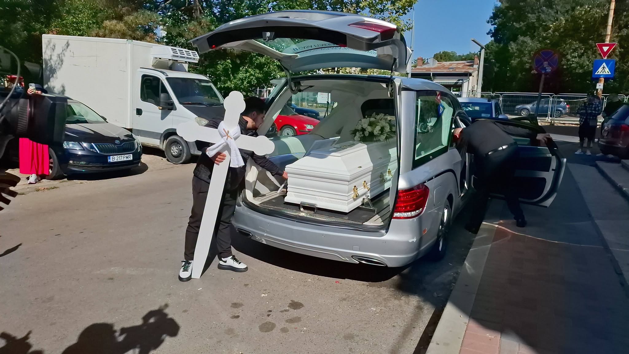 Crucea albă de lemn lângă mașina care a transportat sicriul alb cu trupul Mădălinei Apostol pe ultimul ei drum
