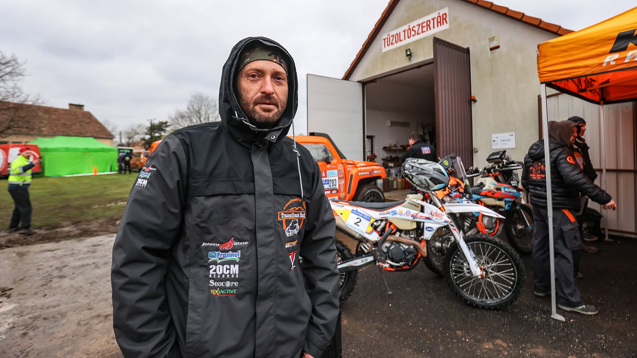 Cheloo &icirc;mbrăcat gros, cu o jachetă groasă cu glugă pe care o ține pe cap, st&acirc;nd l&acirc;ngă mai multe motoare și mașini de teren