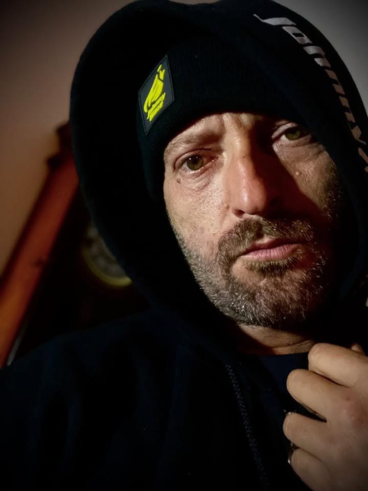 Cheloo făcându-și un selfie într-un hanorac negru, cu căciulă și glugă pe cap, privind serios spre obiectivul camerei