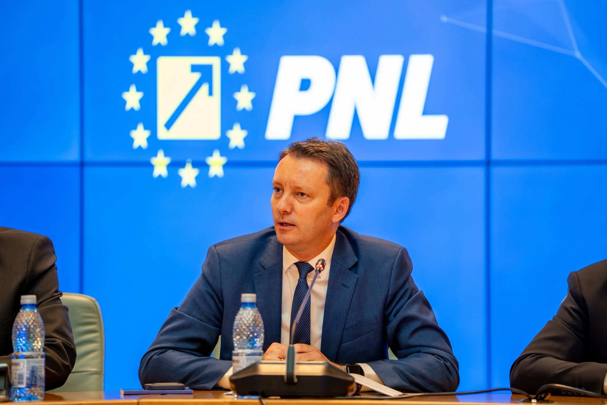 Siegfried Mureșan &icirc;n fața unui ecran pe care este afișată sigla PNL, &icirc;n timp ce era &icirc;mbrăcat &icirc;ntr-un costum bleumarin și vorbea &icirc;n fața altor politicieni