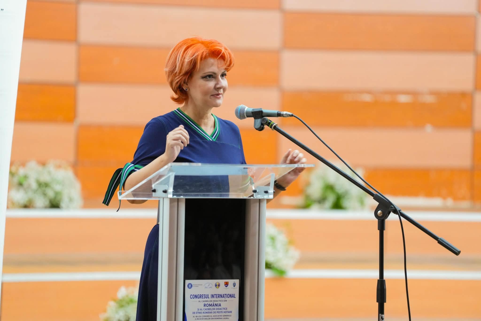 Lia&nbsp;Olguța Vasilescu &icirc;mbrăcată &icirc;n bleumarin, st&acirc;nd la un pupitru și vorbind &icirc;n fața unei audiențe, &icirc;ntr-o sală cu pereți portocalii
