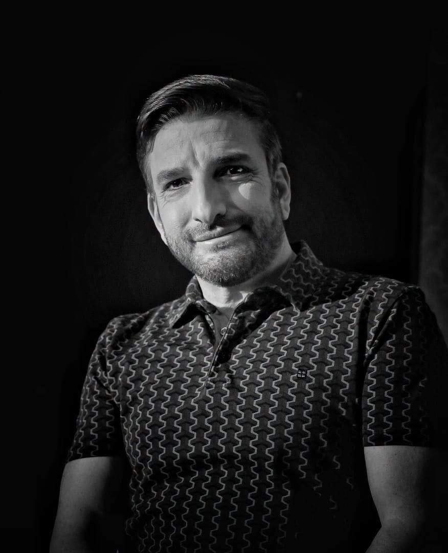 Imagine alb-negru cu Silviu Biriș &icirc;ntr-un tricou &icirc;nchis la culoare cu imprimeuri albe sun formă de coloane
