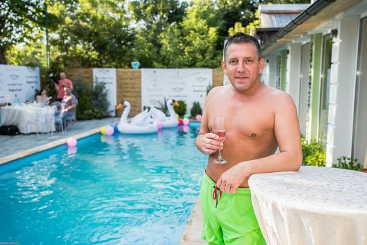 Costin Mărculescu la bustul gol, &icirc;n pantaloni scurți verzi pe marginea unei piscine, sprijinindu-se de o masă și țin&acirc;nd &icirc;n m&acirc;nă un pahar de șampanie