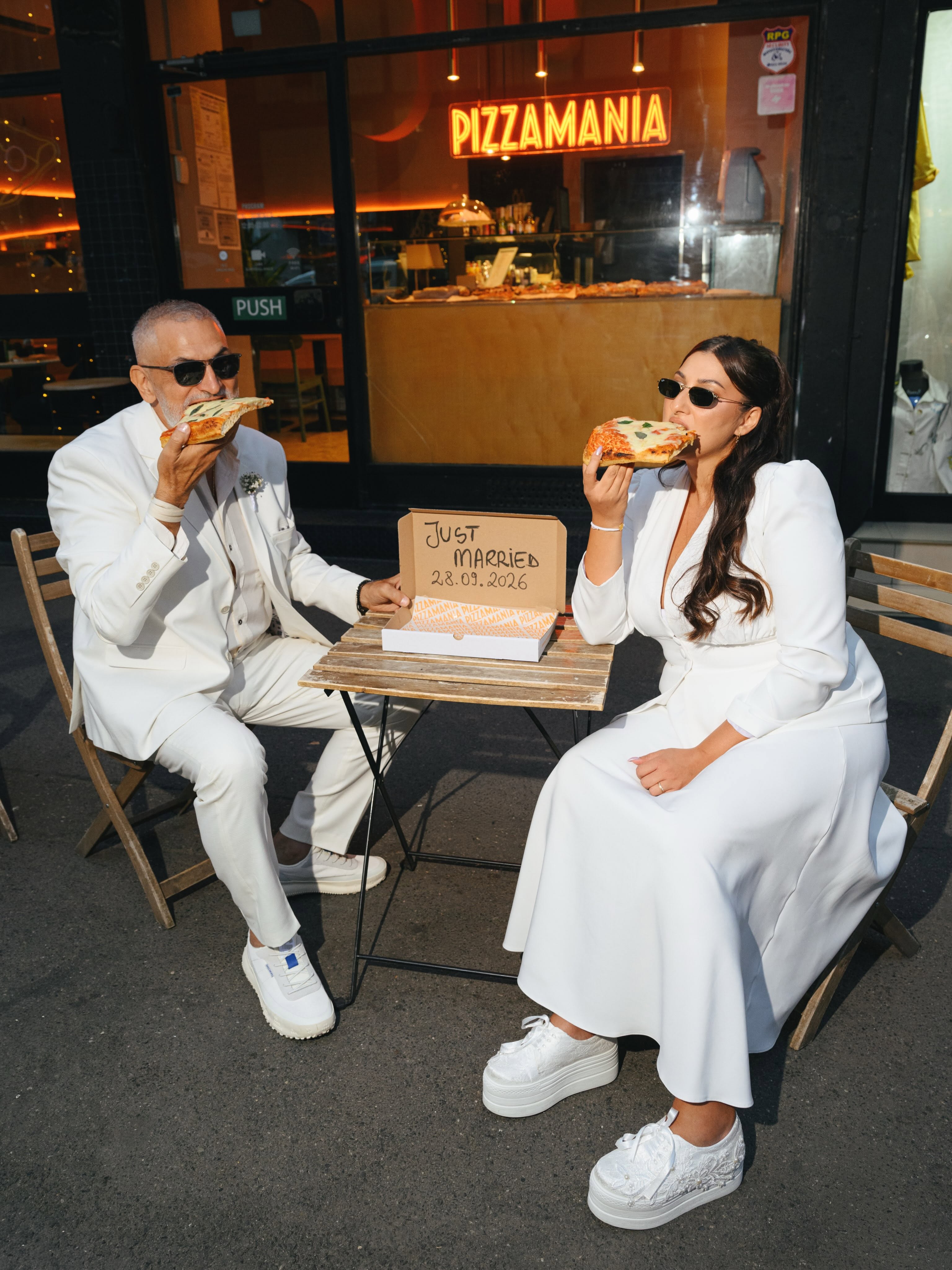 Damian Drăghici și Cristina Stroe m&acirc;nc&acirc;nd pizza după ce s-au căsătorit civil, &icirc;mbrăcați &icirc;n haine albe, cu o cutie de pizza l&acirc;ngă ei pe care este scrisă data căsătoriei