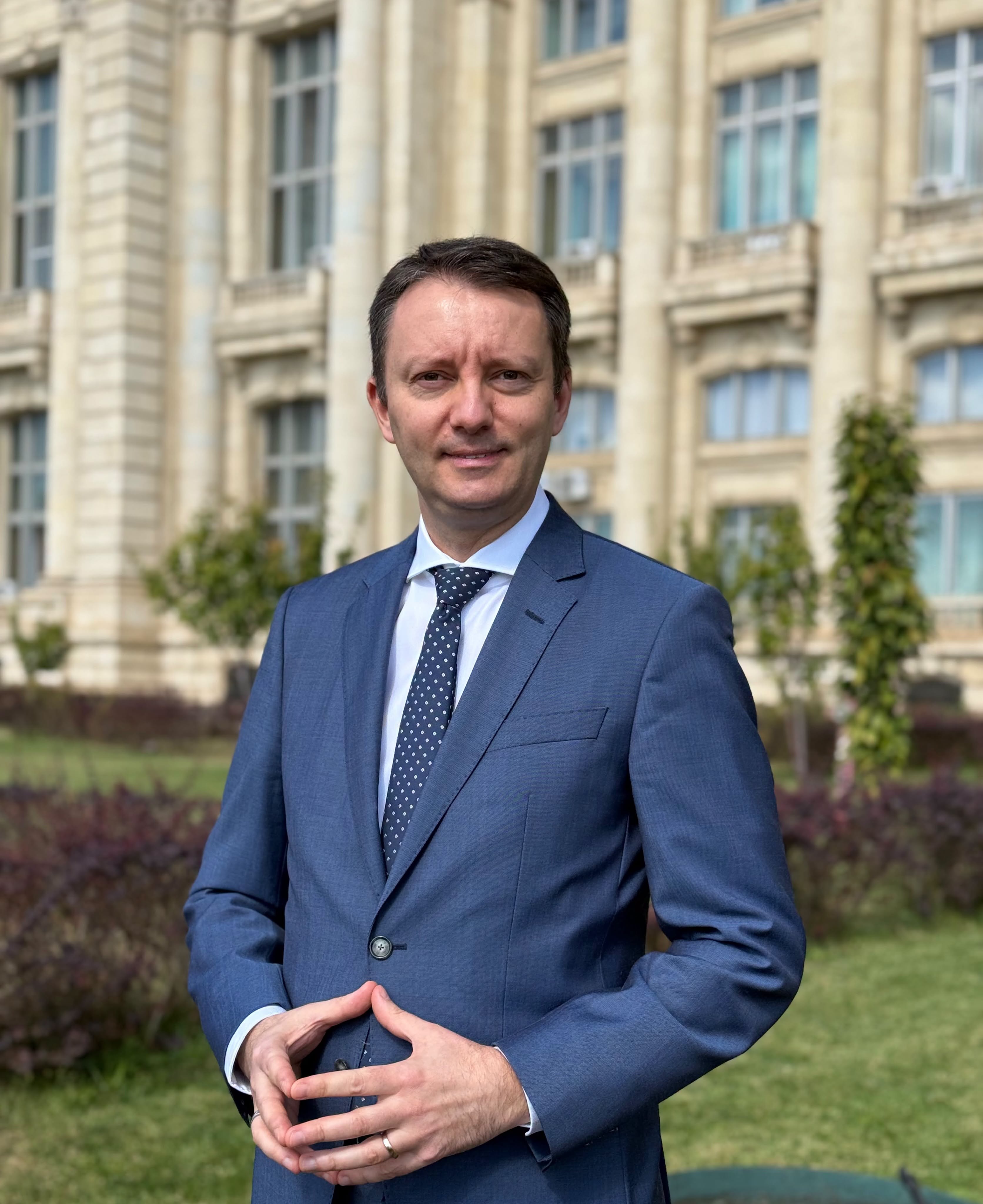 Siegfried Mureșan &icirc;ntr-un costum bleumarin, cu o cravată cu picățele, st&acirc;nd cu m&acirc;inile &icirc;mpreunate &icirc;n fața Parlamentului