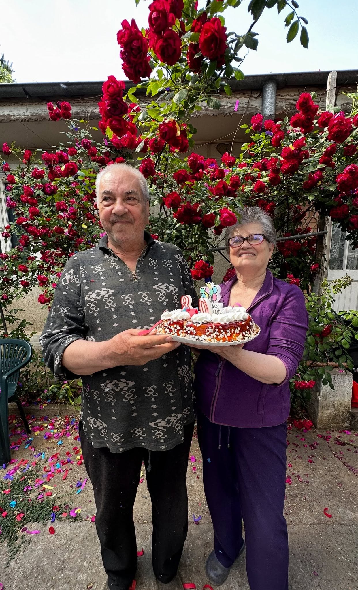 părinții Emmei Ștefan țin&acirc;nd un tort &icirc;n brațe, st&acirc;nd &icirc;n curtea casei, printre flori