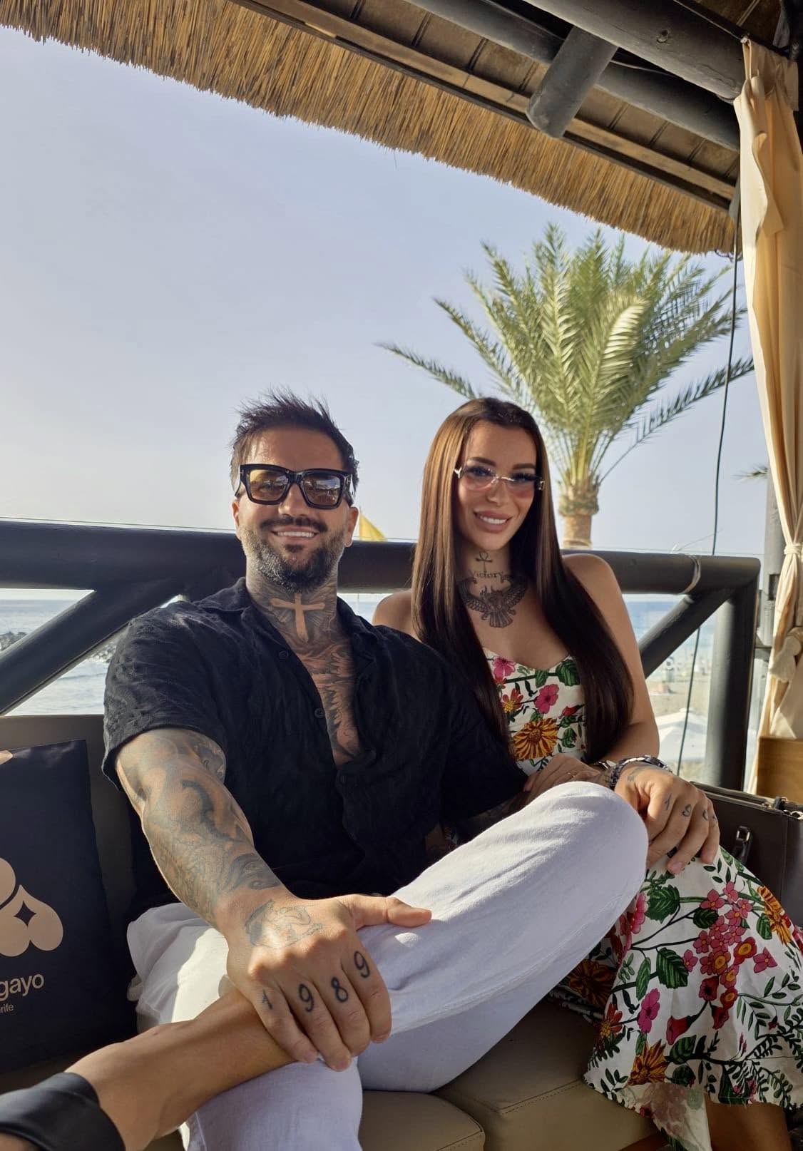 Armina și Marcel Andrei de la &bdquo;Insula Iubirii&rdquo; st&acirc;nd &icirc;mpreună pe canapea, la o terasă cu vedere la mare, purt&acirc;nd ochelari de soare și z&acirc;mbind la camera foto