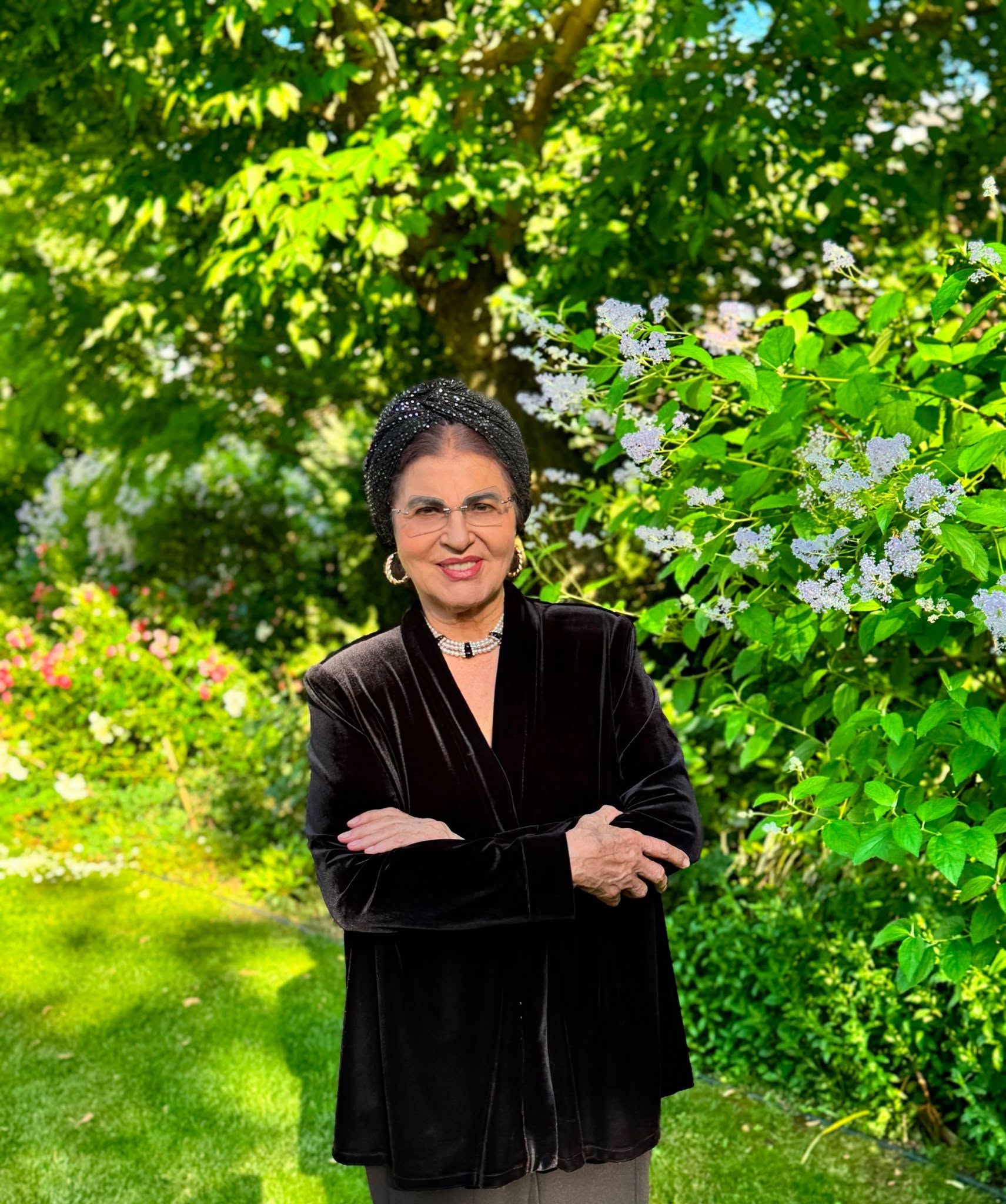 Irina Loghin în grădină, îmbrăcată într-un sacou negru de catifea, ținându-și brațele încrucișate, în timp ce purta pe cap o betiță strălucitoare și la gât un colier din mărgele pe mai multe rânduri