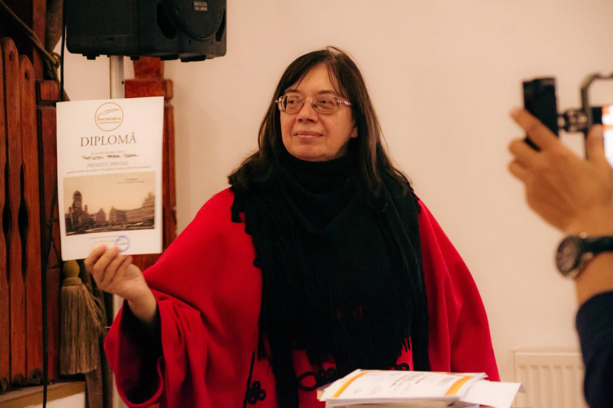 Mihaela Grădinariu z&acirc;mbitoare, &icirc;ntr-un pardesiu roșu, cu o eșarfă neagră, țin&acirc;nd &icirc;n m&acirc;nă o diplomă, &icirc;n cadrul unui eveniment cultural
