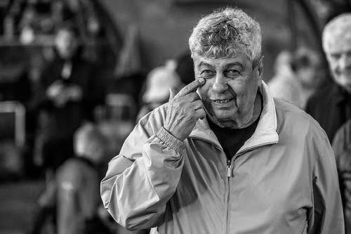 Imagine alb-negru cu Mircea Lucescu z&acirc;mbitor, pe stadion, țin&acirc;ndu-și degetul sub unul dintre ochi