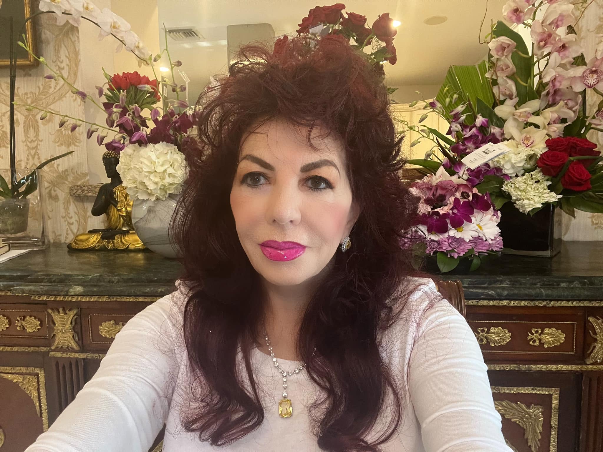 Carmen Harra făc&acirc;ndu-și un selfie l&acirc;ngă o piesă de mobilier pe care sunt așezate foarte multe flori și obiecte decorative