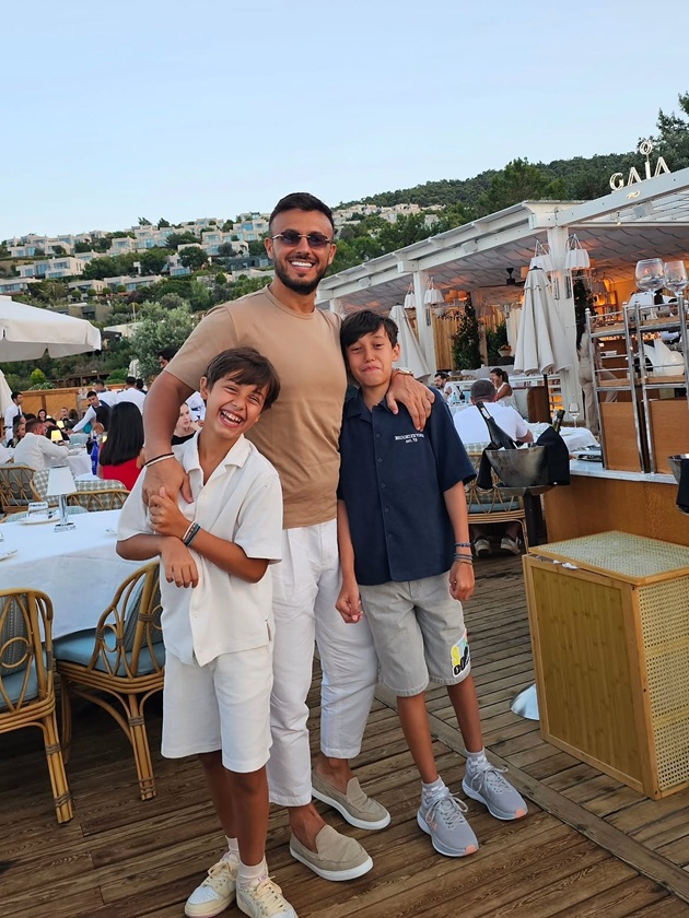 Gabi Bădălău alături de cei doi băieți ai săi care z&acirc;mbesc &icirc;n timp ce el &icirc;i ține de g&acirc;t, &icirc;n timpul unei vacanțe la mare, pe o terasă