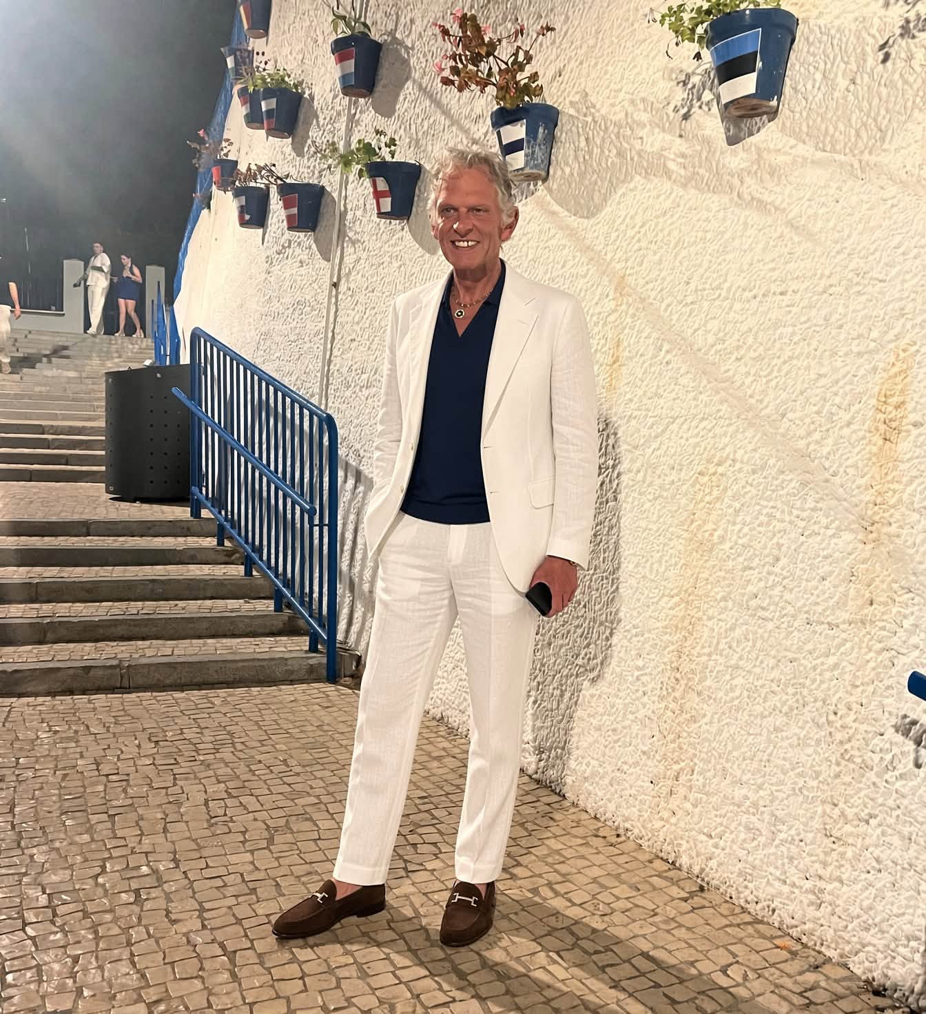Leonard Miron z&acirc;mbitor, &icirc;ntr-un costum alb cu cămașă neagră și pantofi negri, st&acirc;nd l&acirc;ngă un zid alb, plin de ghivece cu flori, la baza unor scări, &icirc;n timpul unei vacanțe