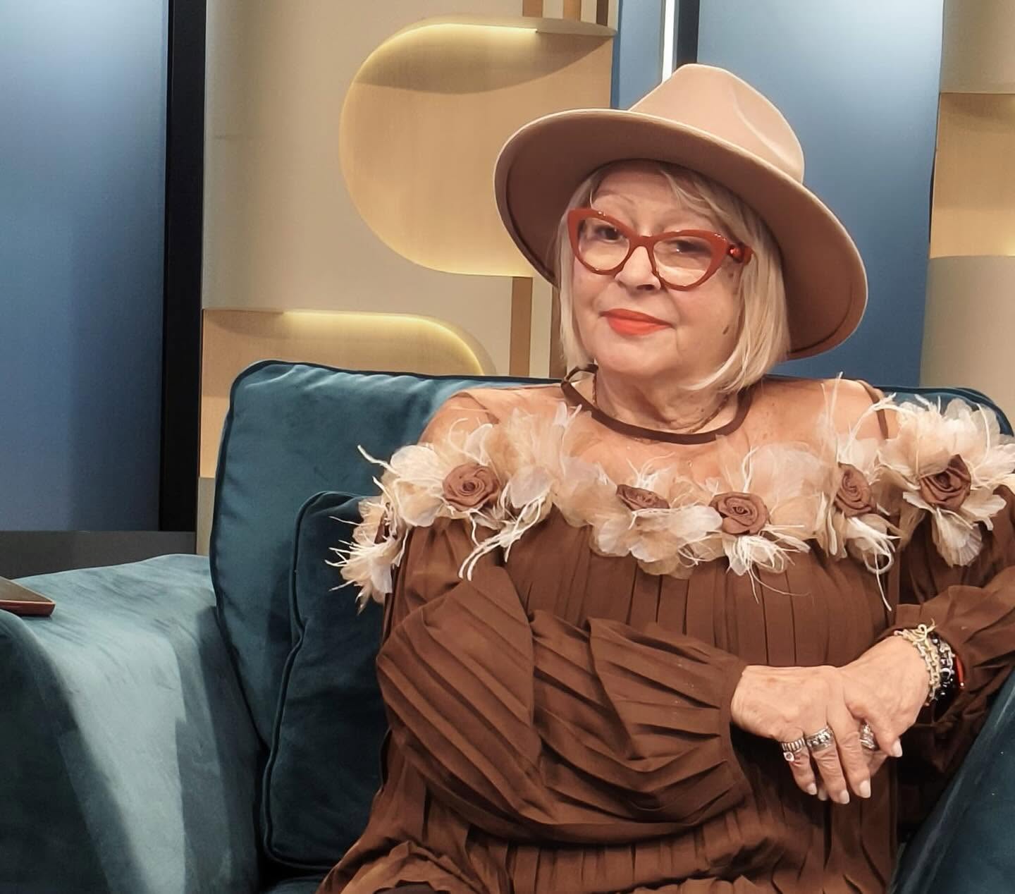 Mirabela Dauer &icirc;ntr-o rochie maro, plisată, cu flori atașate &icirc;n partea de sus și purt&acirc;nd pe cap o pălărie bej
