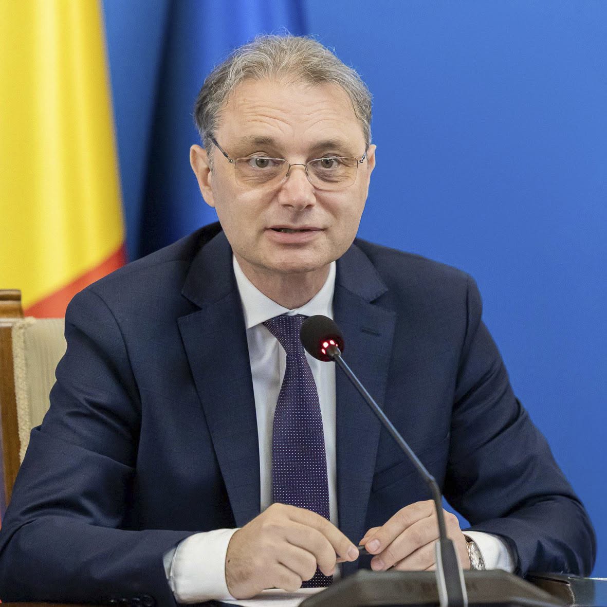 Luca Niculescu, premierul Rom&acirc;niei, st&acirc;nd la birou, cu microfonul &icirc;n față, &icirc;n timp ce ținea &icirc;n m&acirc;nă un pix și avea &icirc;n spate steagul țării