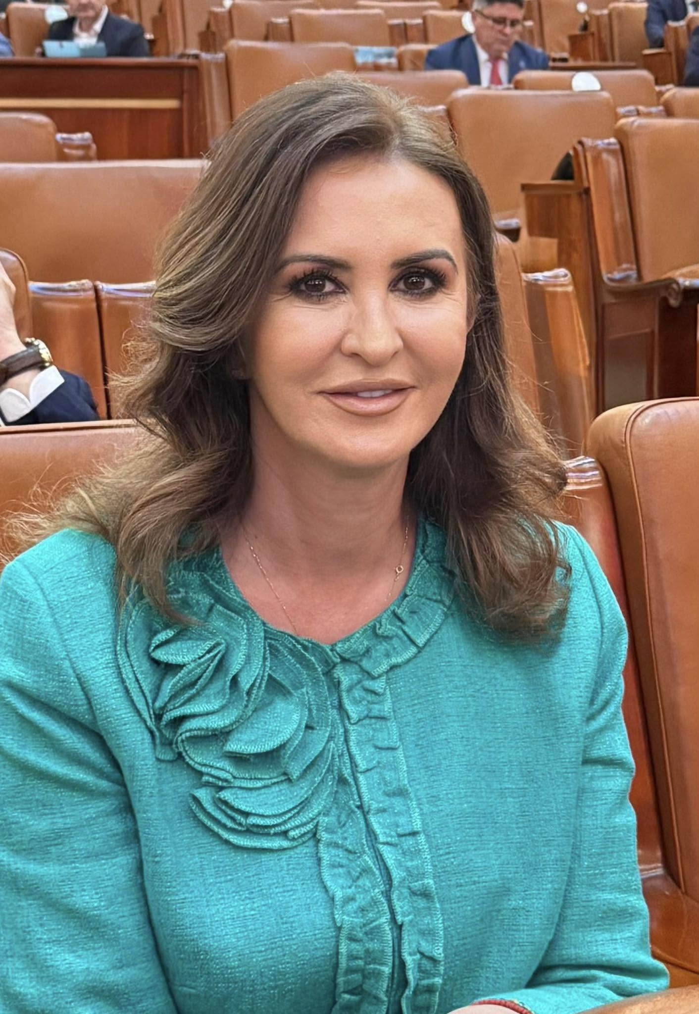 Natalia Intotero, noua ministră a Mediului la Camera Deputaților, st&acirc;nd pe un scaun, &icirc;mbrăcată &icirc;ntr-o bluză turcoaz cu detalii 3D