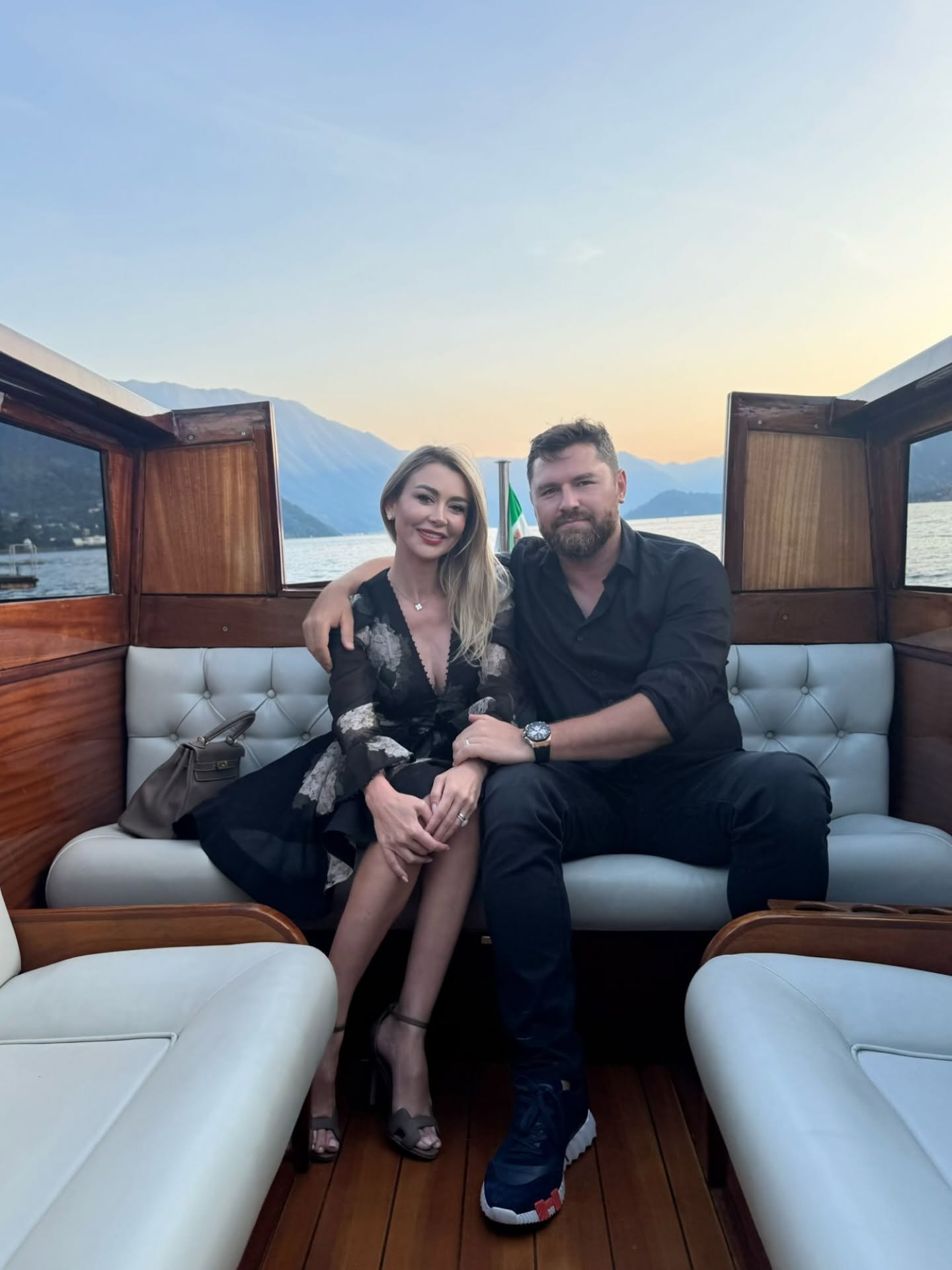 Denisa Tănase și Mircea Br&acirc;nzei pe o ambarcațiune cu scaune și canapele elegante din piele, st&acirc;nd &icirc;mbrățișați și z&acirc;mbitori, av&acirc;nd &icirc;n spate o priveliște superbă