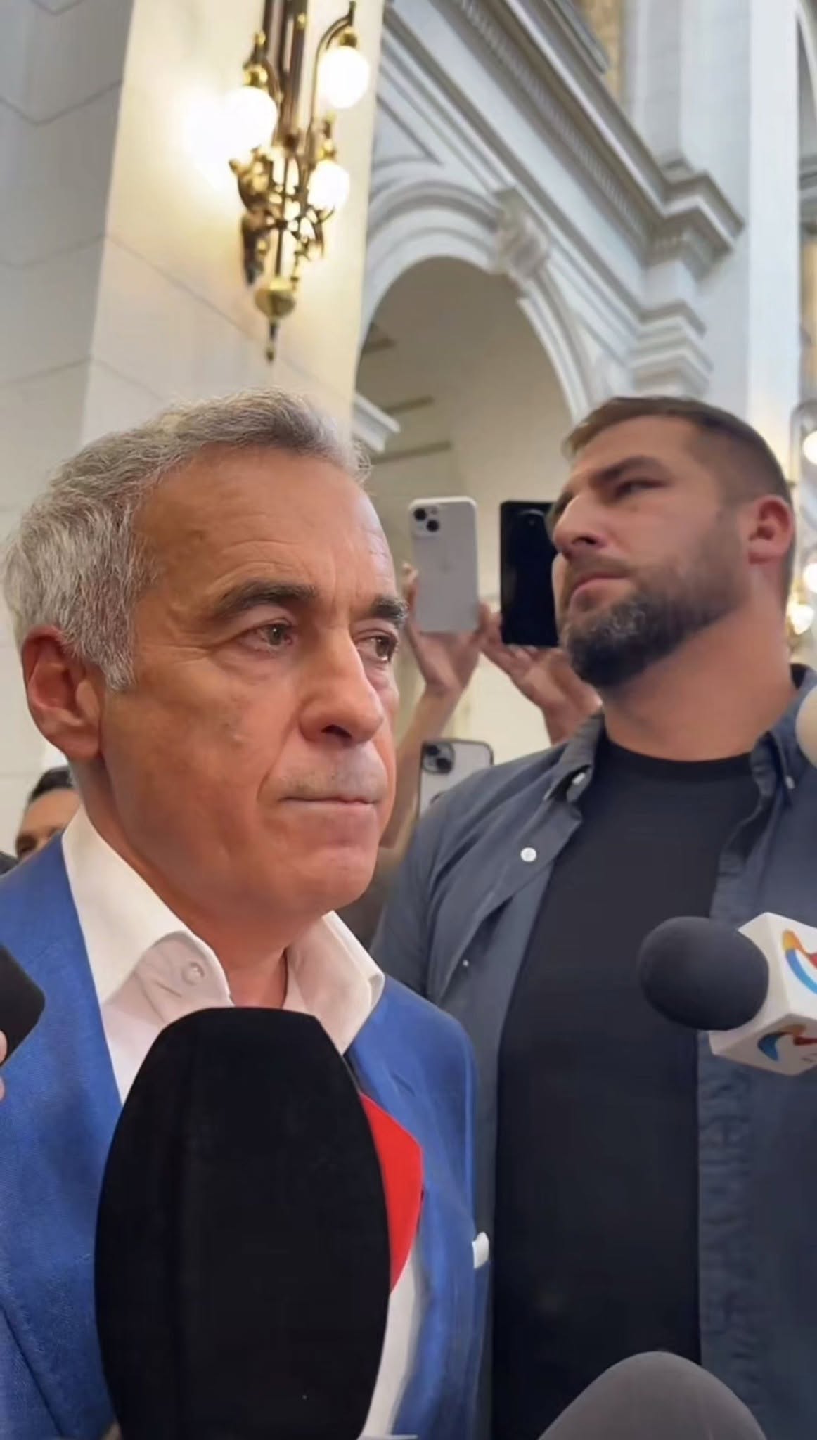 Călin Georgescu înconjurat de oameni și de presă, având în față numeroase microfoane de la diverse posturi TV
